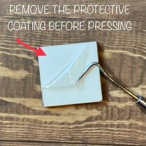 May include: A white ceramic tile with a clear protective film on top. The film is being peeled off with a tool. The text "REMOVE THE PROTECTIVE COATING BEFORE PRESSING" is visible above the tile.