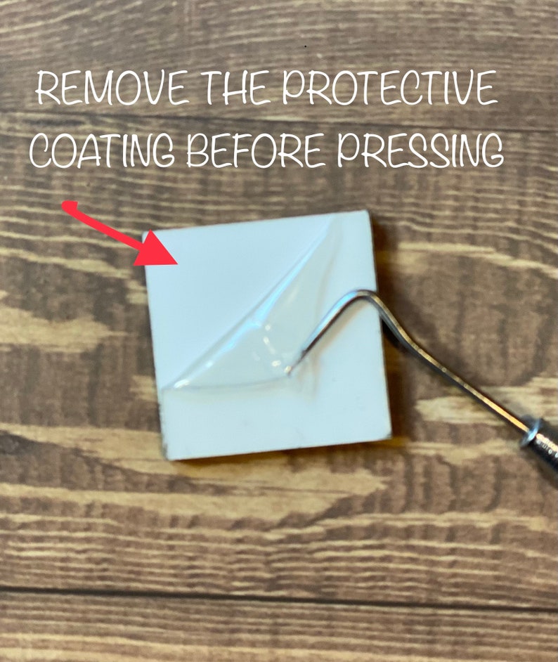 May include: A white ceramic tile with a clear protective film on top. The film is being peeled off with a tool. The text "REMOVE THE PROTECTIVE COATING BEFORE PRESSING" is visible above the tile.