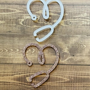 May include: Two stethoscope heart shapes, one white and one with pink glitter. The shapes are made of acrylic and are on a wooden background.