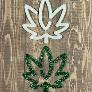 Może przedstawiać: Dwa kształty liści konopi, jeden biały, a drugi zielony z brokatem. Zielony liść znajduje się na białym liściu.