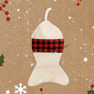 May include: A white stocking with a red and black buffalo plaid cuff. The stocking is hanging against a brown background with white snowflakes and holly.