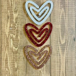 May include: Three layered heart shapes made of wood. The top heart is white, the middle heart is red glitter, and the bottom heart is gold glitter.