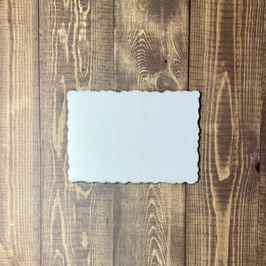 May include: A blank white sign with a scalloped edge, made of wood, against a brown wood background.