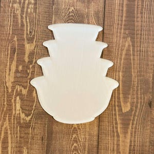 Może przedstawiać: Biały, warstwowy, dekoracyjny przedmiot o unikalnym kształcie, przypominający wielopoziomową misę lub stylizowane naczynie. Obiekt umieszczony jest na brązowym, drewnianym tle, tworząc kontrast kolorów i faktur. Projekt jest prosty, ale przyciągający wzrok.