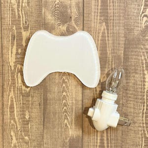 Puede incluir: Una luz de noche blanca con forma de mando, con una bombilla transparente y una base blanca enchufable. La luz de noche está diseñada para parecerse a un mando de videojuegos. Los artículos están sobre una superficie de madera.