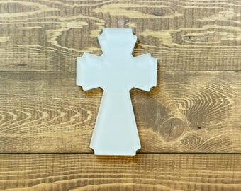 May include: A white, cross-shaped ornament with a textured surface. The cross has a traditional design with a slightly flared base and arms. It is set against a wooden background, creating a contrast in textures and colors.