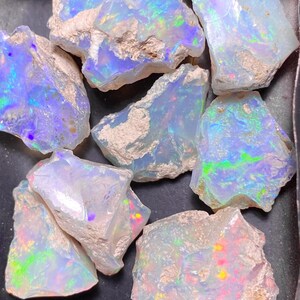 Può includere: Una collezione di opali grezzi con una base bianco latte e lampi iridescenti di blu, verde e giallo. Le pietre preziose grezze sono esposte in un vassoio scuro, mostrando la loro consistenza naturale e il vibrante gioco di colori.
