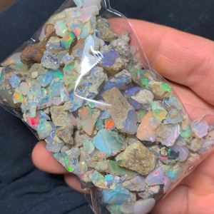 May include: A clear plastic bag filled with rough opal stones. The opals display a variety of colors, including blue, green, red, and orange, with a shimmering, iridescent effect. The stones are a mix of sizes and shapes.