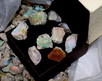 May include: A collection of raw opals in a dark brown box, with a black interior. The opals display a variety of colors, including white, orange, green, and blue. Some opals are also scattered around the box.