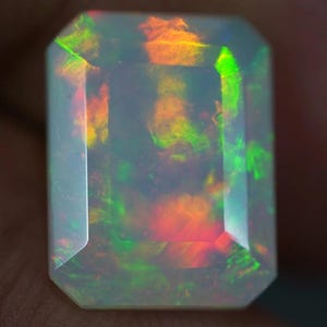May include: A rectangular-cut opal gemstone with a translucent, milky white base. The stone displays vibrant flashes of color, including green, orange, and red, creating an iridescent effect. The gemstone's facets are clearly defined, enhancing its brilliance.