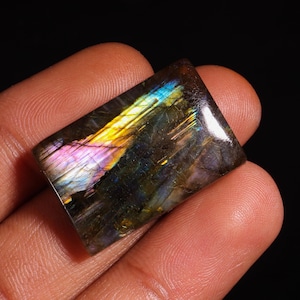 以下が含まれることがあります： 長方形の虹色のラブラドライトの宝石で、濃いグレーのベースに黄色、青、紫の輝きがあります。
