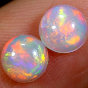 Könnte beinhalten: Zwei runde, irisierende Opal-Edelsteine mit einer weißen Basis und Blitzen von Orange, Rot, Grün und Blau.