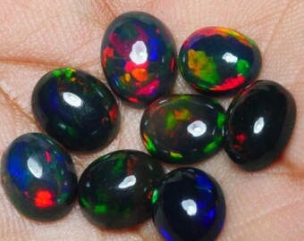Puede incluir: Siete opales negras de forma ovalada con destellos iridiscentes verdes, azules y rojos. Las opales están sobre una superficie marrón claro.