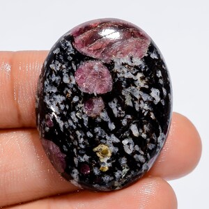 Może przedstawiać: Owalny kamień szlachetny w szlifie kaboszonowym z czarno-białym, nakrapianym tłem i różowymi wrostkami rubinu.