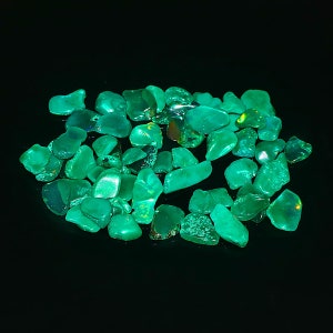 May include: A collection of green gemstone chips, likely opal, on a black background. The stones are various shapes and sizes, with some showing a slight iridescent sheen.