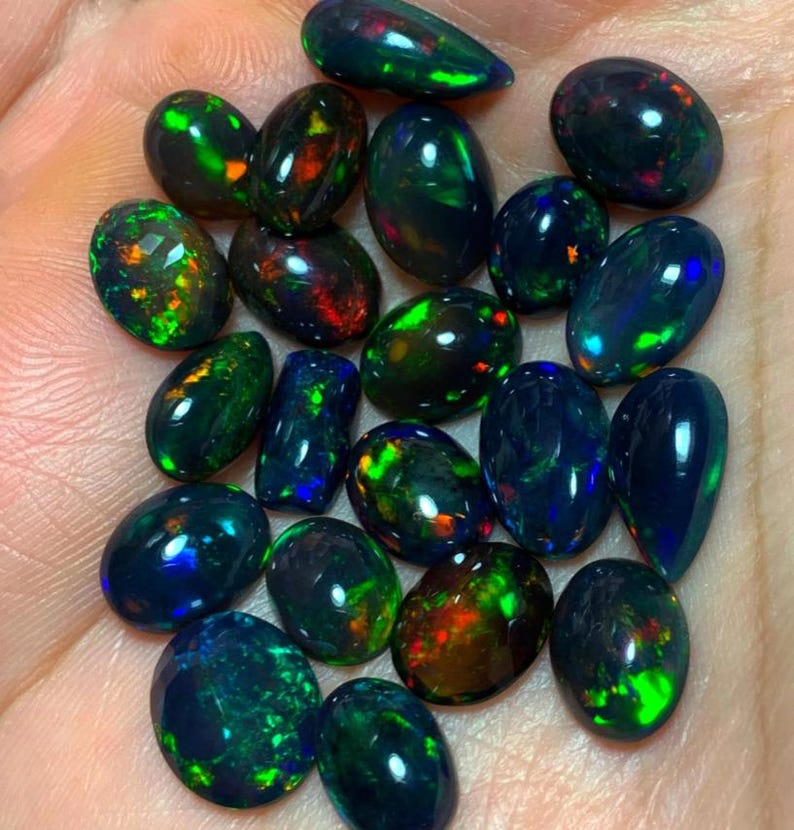 May include: A collection of black opal gemstones in various shades of green, blue, and red. The opals are pear-shaped and have a smooth, polished finish.