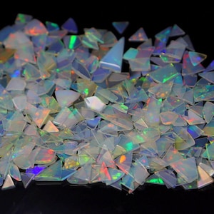 Peut inclure: Un tas d'éclats d'opale iridescents, coupés en diverses formes géométriques, dont des triangles et des formes irrégulières. Les opales présentent un jeu de couleurs avec des éclats de vert, de bleu et de rouge, sur fond noir.