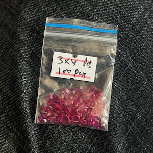 May include: A small clear plastic bag with a blue stripe at the top, containing numerous small, faceted, pink gemstones. A handwritten label reads "3x4 ps 100 per". The bag is set against a dark, textured background.