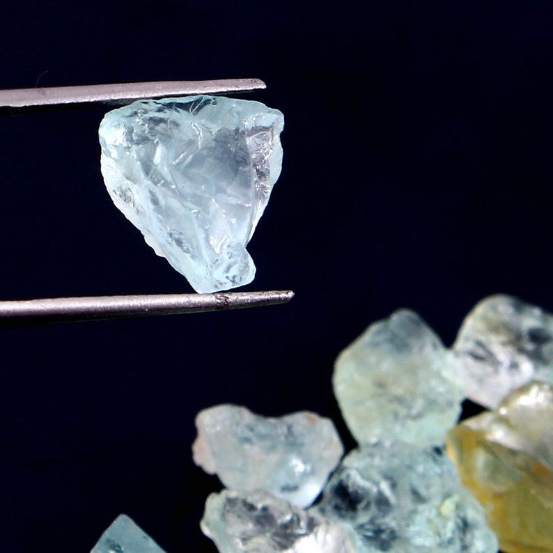 May include: A close-up of a light blue aquamarine crystal held by tweezers. The crystal is faceted and has a translucent appearance. Several other aquamarine crystals are visible in the background.