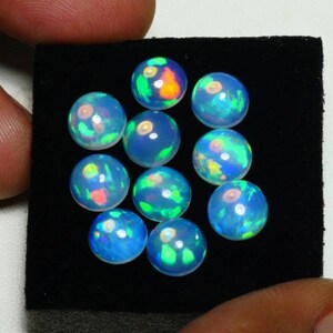 May include: A collection of 10 round, iridescent opal cabochons in various shades of blue, green, and white. The cabochons are displayed on a black velvet background.
