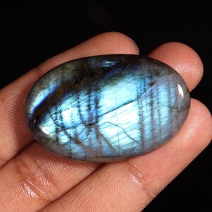Peut inclure: Une pierre précieuse de labradorite de forme ovale avec une surface polie. La pierre présente un jeu de couleurs saisissant, avec des éclairs irisés de bleu et de turquoise sur une base gris foncé. La pierre est tenue dans une main sur fond noir.