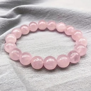 May include: A pink rose quartz beaded bracelet with round beads. The bracelet is on a white fabric background.