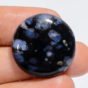 Könnte beinhalten: Ein runder, schwarzer und blauer Edelstein-Cabochon mit einem gesprenkelten Muster. Der Edelstein hat eine glatte, polierte Oberfläche.