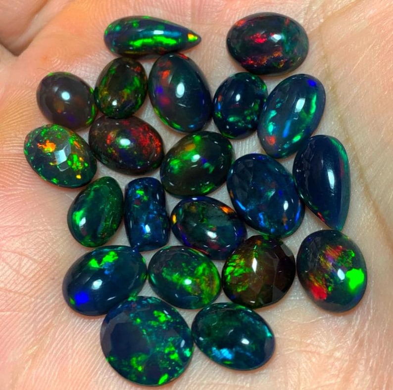 May include: A collection of black opal gemstones with green, blue, and red iridescent flashes. The opals are oval and teardrop shaped.