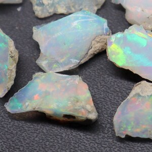 以下が含まれることがあります： 虹色のオパール原石のコレクション。石は様々な形とサイズで、乳白色のベースに緑、青、オレンジの輝きがあります。