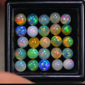 Könnte beinhalten: Eine Sammlung von 25 runden Opal-Edelsteinen in verschiedenen Farben, darunter Grün, Blau, Orange und Weiß, die in einer schwarzen quadratischen Schachtel präsentiert werden.