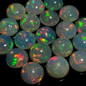 Könnte beinhalten: Eine Sammlung von 18 runden, irisierenden Opal-Cabochons. Die Opale sind in einem blassen Weiß mit Schimmern in Grün, Blau, Rot und Orange.