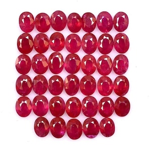 May include: A collection of oval-cut, deep red gemstones. The gemstones are arranged closely together, filling the frame. The polished stones have a high luster, reflecting light. These rubies are suitable for jewelry making.
