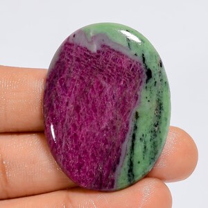May include: Oval-shaped ruby zoisite gemstone. The stone features a vibrant mix of colors, with a deep magenta center and a green border. The polished surface reflects light, highlighting the natural patterns and textures.