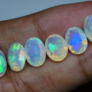 May include: Six oval-shaped opals with iridescent colors, including green, blue, and orange, are displayed on a hand.