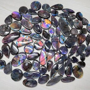 May include: A collection of polished labradorite gemstones in various shapes, including ovals, teardrops, and rounds. The stones display iridescent flashes of blue, purple, and gold, set against a dark gray base. The gemstones are arranged on a light-colored surface.