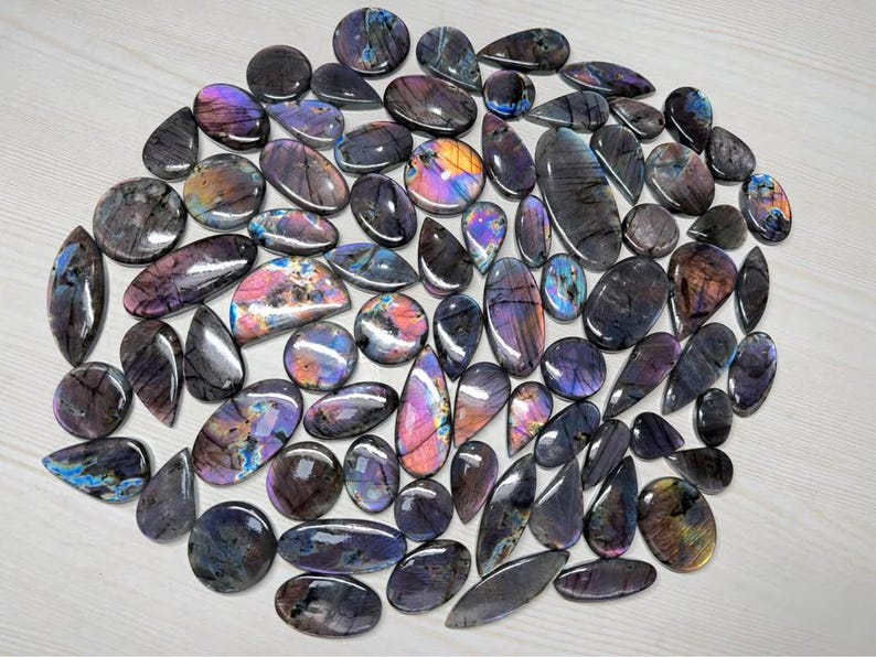 May include: A collection of polished labradorite gemstones in various shapes, including ovals, teardrops, and rounds. The stones display iridescent flashes of blue, purple, and gold, set against a dark gray base. The gemstones are arranged on a light-colored surface.