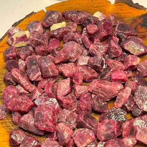 Pode incluir: Uma coleção de rubis brutos e não polidos em vários tons de rosa profundo e vermelho escuro. As pedras têm formas irregulares e irregulares com superfícies texturizadas, com algumas peças apresentando pequenas inclusões de minerais brancos.