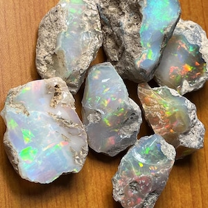 May include: A collection of rough opal stones with iridescent colors, including green, blue, and red. The stones are in various shapes and sizes and are set against a wooden background.