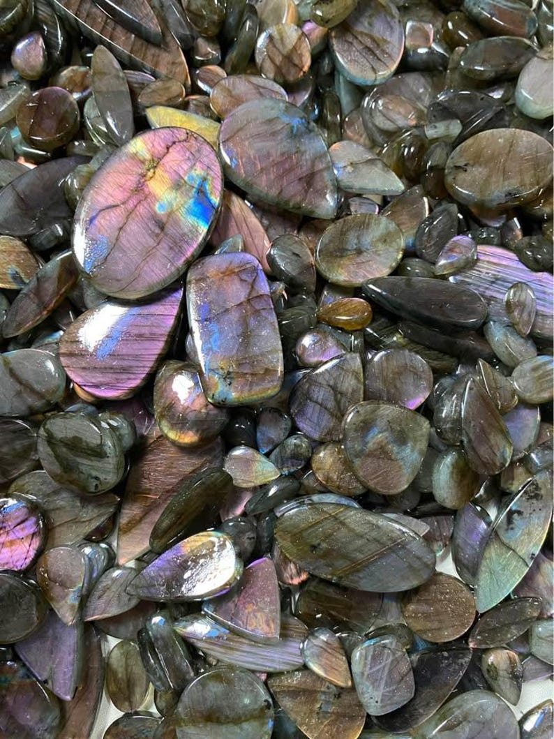 May include: A collection of polished labradorite gemstones in various shapes and sizes. The stones display a range of colors, including purple, blue, and green, with iridescent flashes. The gemstones are oval, teardrop, and irregular shapes.