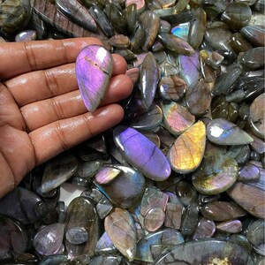 May include: A collection of polished labradorite gemstones in various shapes and sizes. The stones display iridescent flashes of purple, blue, and gold. One stone is held in a hand, showcasing its vibrant color and unique pattern. The gemstones are a mix of oval, teardrop, and irregular shapes.