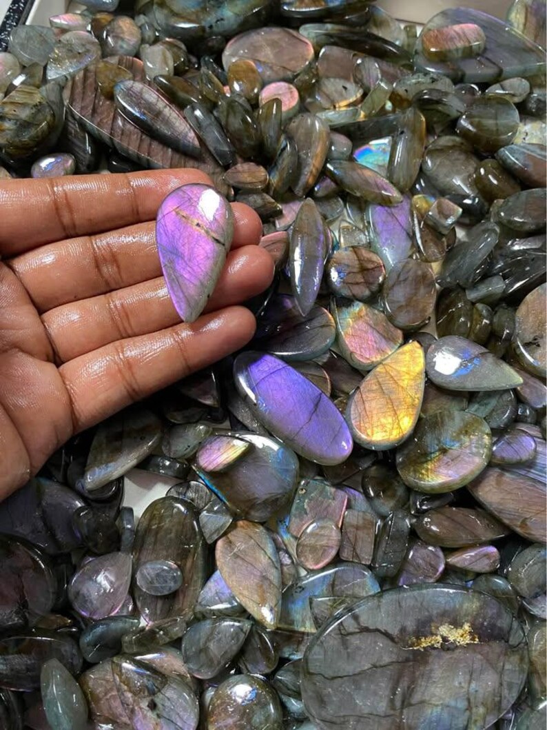May include: A collection of polished labradorite gemstones in various shapes and sizes. The stones display iridescent flashes of purple, blue, and gold. One stone is held in a hand, showcasing its vibrant color and unique pattern. The gemstones are a mix of oval, teardrop, and irregular shapes.