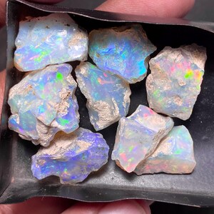 May include: A collection of rough opal gemstones, showcasing a play of color with iridescent flashes of blue, green, and red. The opals are in various sizes and shapes, with a raw, natural texture. The gemstones are displayed in a dark tray.