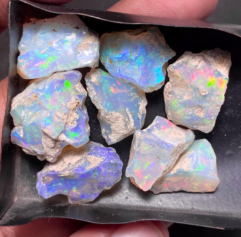 May include: A collection of rough opal gemstones, showcasing a play of color with iridescent flashes of blue, green, and red. The opals are in various sizes and shapes, with a raw, natural texture. The gemstones are displayed in a dark tray.