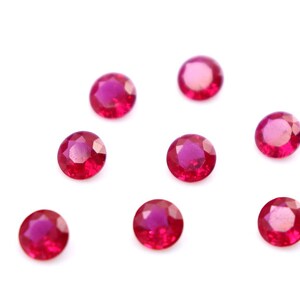 May include: A collection of seven round, faceted, deep red gemstones, possibly rubies, are scattered on a white background. The gems have a polished, reflective surface, and their color is consistent throughout.