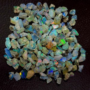 Puede incluir: Una colección de piedras preciosas de ópalo en bruto. Los ópalos muestran una variedad de colores, incluyendo azul, verde y destellos de tonos iridiscentes. Las piedras en bruto tienen una apariencia natural, sin pulir, con una mezcla de tamaños y formas.