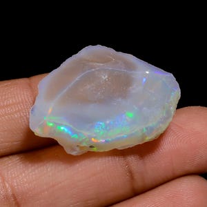 Puede incluir: Un ópalo en bruto y translúcido con una forma irregular y redondeada. La piedra presenta un color base blanco lechoso con destellos vibrantes de luz iridiscente verde, azul y naranja que se reflejan en su superficie cristalina y texturizada.