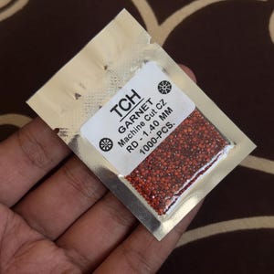 Pode incluir: Um pequeno pacote de folha de alumínio prateada contendo numerosas pedras preciosas de granada castanho-avermelhadas. O pacote tem um rótulo branco com o texto "TCH GARNET Machine Cut CZ RD - 1,40 MM 1000-PCS."