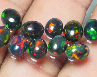 Puede incluir: Una colección de diez ópalos negros ovalados. Cada piedra muestra destellos vibrantes de color, incluyendo rojo, verde y naranja, sobre un fondo oscuro. Los ópalos son lisos y pulidos, reflejando la luz.