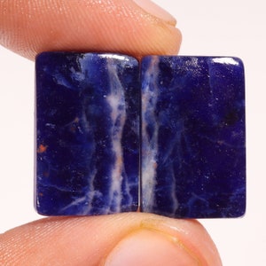 May include: Two rectangular sodalite gemstones. The stones are a deep blue color with white and orange veining. The gemstones are held between fingers, showcasing their polished surfaces and natural patterns. These stones are ideal for jewelry making.