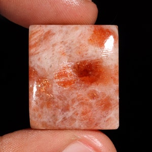 May include: A square, polished sunstone cabochon with a reddish-orange and white color pattern. The stone has a translucent appearance with a slight shimmer.